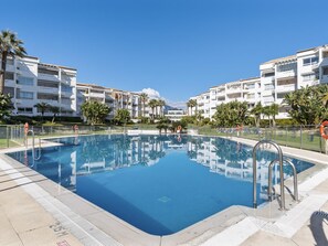 A swimming pool surrounded by multi-story buildings with balconies, clear skies, and palm trees. - Apartamento en Playa Roc o (Marbella)