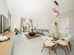 Ein modernes Wohnzimmer mit einer Couch, einem Esstisch mit Stühlen, einem Fernseher und einem Balkon mit Vorhängen.