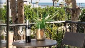 Uma varanda com mesa, uma garrafa e uma planta, com vista para uma praia com palmeiras e o mar.