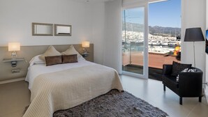 Ett hotellrum med en säng, två sänglampor, en soffa, ett stort fönster med utsikt över en småbåtshamn och en balkong.