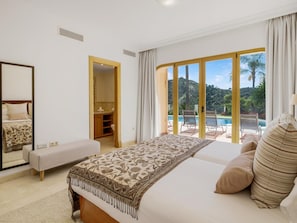 A bedroom with a large bed, a bench, a bathroom, and a view of a pool and greenery. - Villa Los Arqueros Golf (Benahavis)