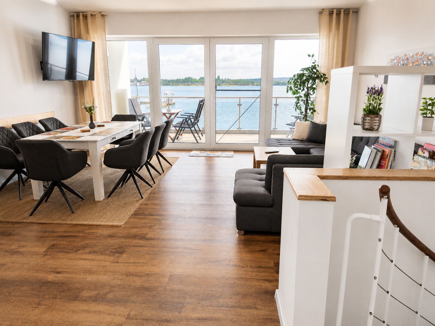 A modern living room with a dining area, a sofa, a TV, and a view of a body of water.
