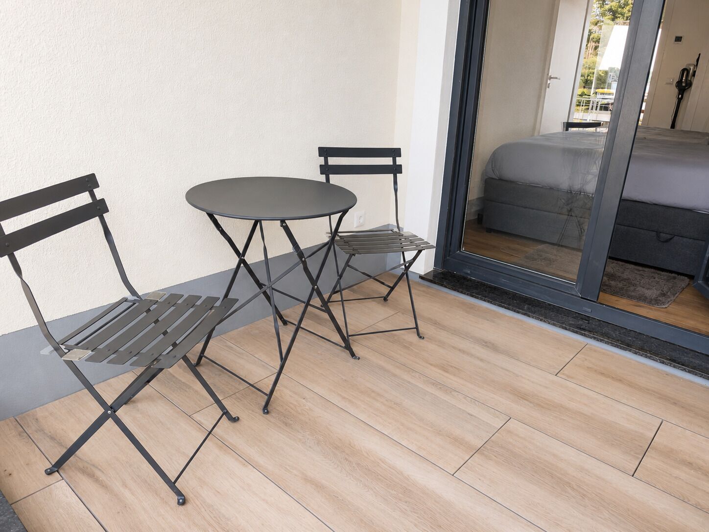 Ein kleiner Patio-Bereich im Freien mit einem runden Tisch und zwei Stühlen, einer Glasschiebetür, die zu einem Zimmer führt, und einem Holzboden.