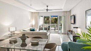 A modern living room with a glass dining table, sofa, and chairs, a ceiling fan, and a view through a glass door to an outdoor patio. - Apartamento en Playas del Duque ref 102 (Marbella)