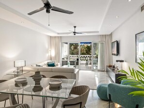 A modern living room with a glass dining table, sofa, and chairs, a ceiling fan, and a view through a glass door to an outdoor patio. - Apartamento en Playas del Duque ref 102 (Marbella)