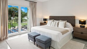 A bedroom with a large bed, two bedside tables, a nightstand, a window with curtains, and a view of a pool and greenery. - Apartamento en Playas del Duque ref 102 (Marbella)