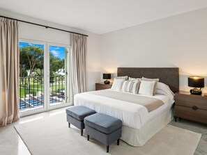 A bedroom with a large bed, two bedside tables, a nightstand, a window with curtains, and a view of a pool and greenery. - Apartamento en Playas del Duque ref 102 (Marbella)