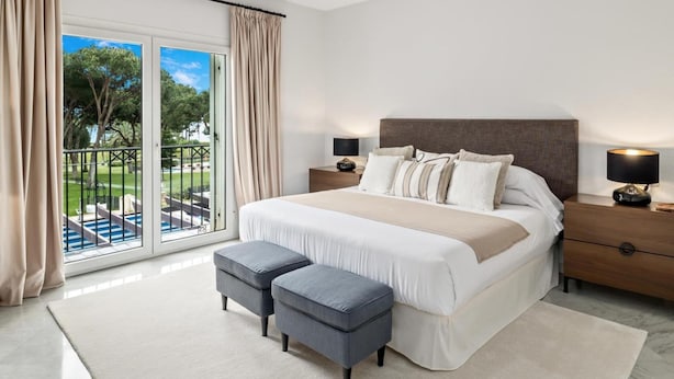 A bedroom with a large bed, two bedside tables, a nightstand, a balcony with curtains, and a view of a pool and greenery. - Apartamento en Playas del Duque ref 102 (Marbella)
