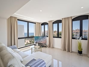 Ein modernes Wohnzimmer mit einer Couch, einem Couchtisch und einem Gemälde. Große Fenster bieten Blick auf die Stadt.