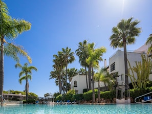 A pool area with clear blue water, surrounded by palm trees and a white building with balconies. - Jardines de Aldaba 412 (Marbella)
