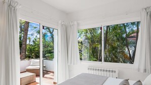 Ein Schlafzimmer mit Bett, Heizkörper und Blick auf einen Garten durch große Fenster.