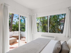 A bedroom with a bed, a radiator, and a view of a garden through large windows. - Jardines de Aldaba 412 (Marbella)