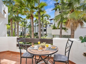 A round table with chairs, a breakfast setting, and a view of palm trees and buildings. - Jardines de Aldaba 412 (Marbella)
