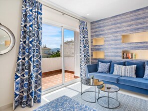 Ein modernes Wohnzimmer mit einem blauen Sofa, gemusterten Vorhängen, einer Glastür zu einem Balkon und einer wandmontierten Bücherreolen.
