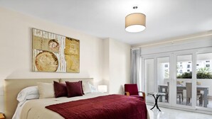 A hotel room with a large bed, a painting on the wall, a view of the outside through a glass door, and a small table with chairs. - Amplio Apartamento en Playas del Duque (Marbella)