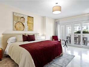 A hotel room with a large bed, a painting on the wall, a view of the outside through a glass door, and a small table with chairs. - Amplio Apartamento en Playas del Duque (Marbella)