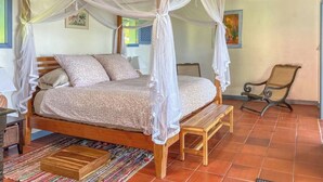 A four-poster canopy bed with a bench beside it, a wooden bench, a chair, and a colorful woven rug.