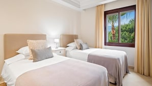 A hotel room with two beds, a nightstand, a window with a view of greenery, and beige curtains. - Apartamento Junto al mar en Estepona (Estepona)