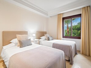 A hotel room with two beds, a nightstand, a window with a view of greenery, and beige curtains. - Apartamento Junto al mar en Estepona (Estepona)