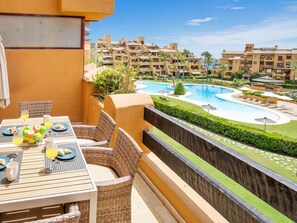 A balcony with wicker furniture, a dining table set with plates, glasses, and a fruit bowl. In the background, there is a swimming pool surrounded by greenery and a row of buildings. - Apartamento Junto al mar en Estepona (Estepona)