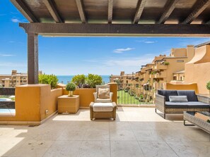 A covered outdoor seating area with wicker furniture, a small table with a plant, and a view of a coastal town. - Apartamento Junto al mar en Estepona (Estepona)