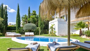 A pool area with lounge chairs, a thatched umbrella, and a white building in the background. - Preciosa Villa con Magn Ficas Vistas en Los Flamin (Benahavis)