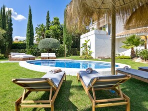 A pool area with lounge chairs, a thatched umbrella, and a white building in the background. - Preciosa Villa con Magn Ficas Vistas en Los Flamin (Benahavis)