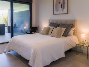 Ein modernes Schlafzimmer mit einem großen Bett, einer Glastür zu einem Außenbereich, einem Nachttisch mit einer Lampe und einem gerahmten abstrakten Gemälde an der Wand.