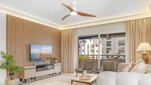 A modern living room with a flat-screen TV, comfortable seating, a ceiling fan, and a view of the city through large windows. - Marina Ban s c Ntrico Apartamento (Marbella)