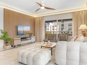 A modern living room with a flat-screen TV, comfortable seating, a ceiling fan, and a view of the city through large windows. - Marina Ban s c Ntrico Apartamento (Marbella)