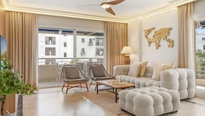 A modern living room with a sofa, two armchairs, a coffee table, a ceiling fan, and a view of a building through large windows. - Marina Ban s c Ntrico Apartamento (Marbella)