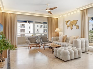 A modern living room with a sofa, two armchairs, a coffee table, a ceiling fan, and a view of a building through large windows. - Marina Ban s c Ntrico Apartamento (Marbella)
