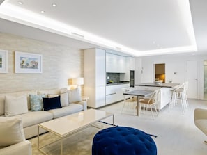 Ein modernes Wohnzimmer mit Sofa, Couchtisch und Hocker, ein Essbereich mit Stühlen und Tisch sowie eine Küche mit Schränken und Geräten.