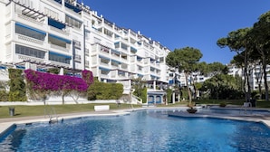 A swimming pool with a clear blue water surface, surrounded by a white building with balconies, and a tree with purple flowers to the left. - Amplio y Luminoso Apartamento en 1 l nea Playa (Marbella)