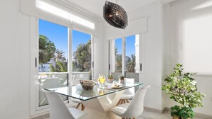 A modern dining area with a glass table, white chairs, and a view of palm trees through large windows. - Amplio y Luminoso Apartamento en 1 l nea Playa (Marbella)