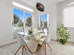 Ein moderner Essbereich mit einem Glastisch, weißen Stühlen und Blick auf Palmen durch große Fenster.