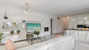 A modern living room with a white sofa, a glass coffee table, and a TV. There is a kitchen area with white cabinets and a bar counter. - Amplio y Luminoso Apartamento en 1 l nea Playa (Marbella)