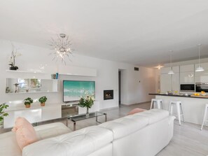 Ein modernes Wohnzimmer mit einem weißen Sofa, einem Glastisch und einem Fernseher. Es gibt eine Küchenzeile mit weißen Schränken und einer Theke.