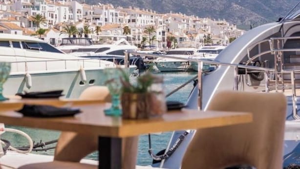 A marina with boats docked, a table with chairs, and a view of coastal buildings. - Stunning Marina Views in Puerto Banus (Marbella)