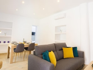 Ein modernes Wohnzimmer mit einem grauen Sofa, einem Esstisch mit Stühlen und einer durch eine offene Tür einsehbaren Küche.