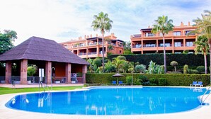 Uma piscina com um pavilhão de telhado de palha, rodeada por sebes bem cuidadas e palmeiras, com edifícios de vários andares ao fundo.