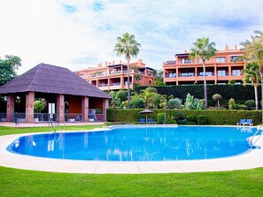 Ein Swimmingpool mit einem strohgedeckten Pavillon, umgeben von gepflegten Hecken und Palmen, mit mehrstöckigen Gebäuden im Hintergrund.