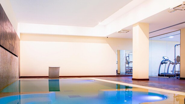 Piscina interna com água cristalina, piso de madeira e equipamentos de ginástica ao fundo.
