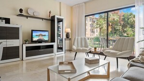 A modern living room with a flat-screen TV, a sofa, two armchairs, a coffee table, and a balcony with a view of palm trees and buildings. - Gran Terraza ref ADM 116 (Marbella)