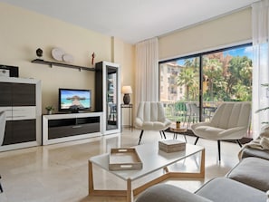 Ein modernes Wohnzimmer mit Flachbildfernseher, einer Couch, zwei Sesseln, einem Couchtisch und einem Balkon mit Blick auf Palmen und Gebäude.