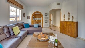 A living room with a sofa, a coffee table with a bottle and glass, and a view into the kitchen. - Contemporaly Huge Riyad (Estepona)