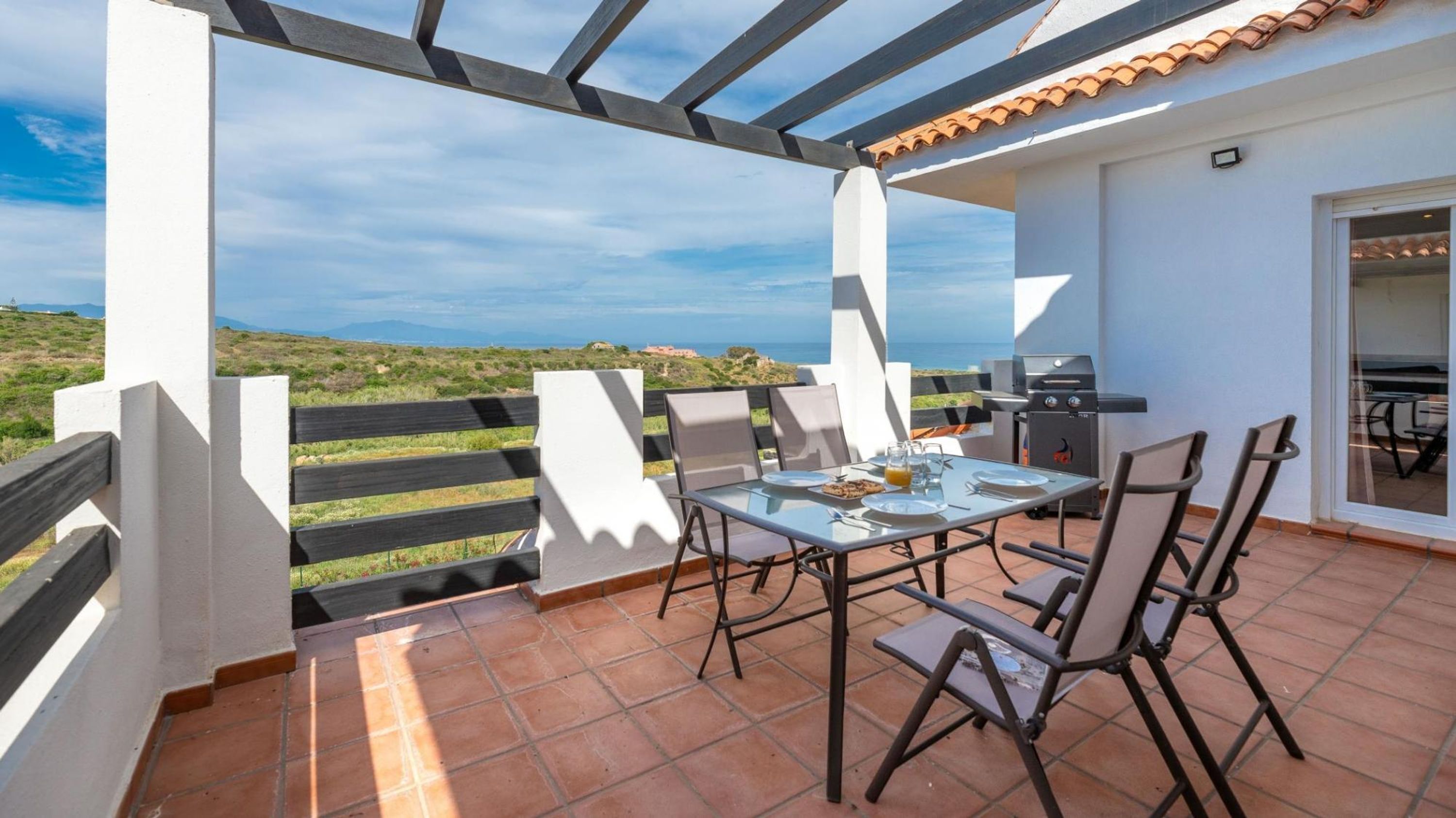 An outdoor patio with a table and chairs, a grill, and a view of the landscape.
