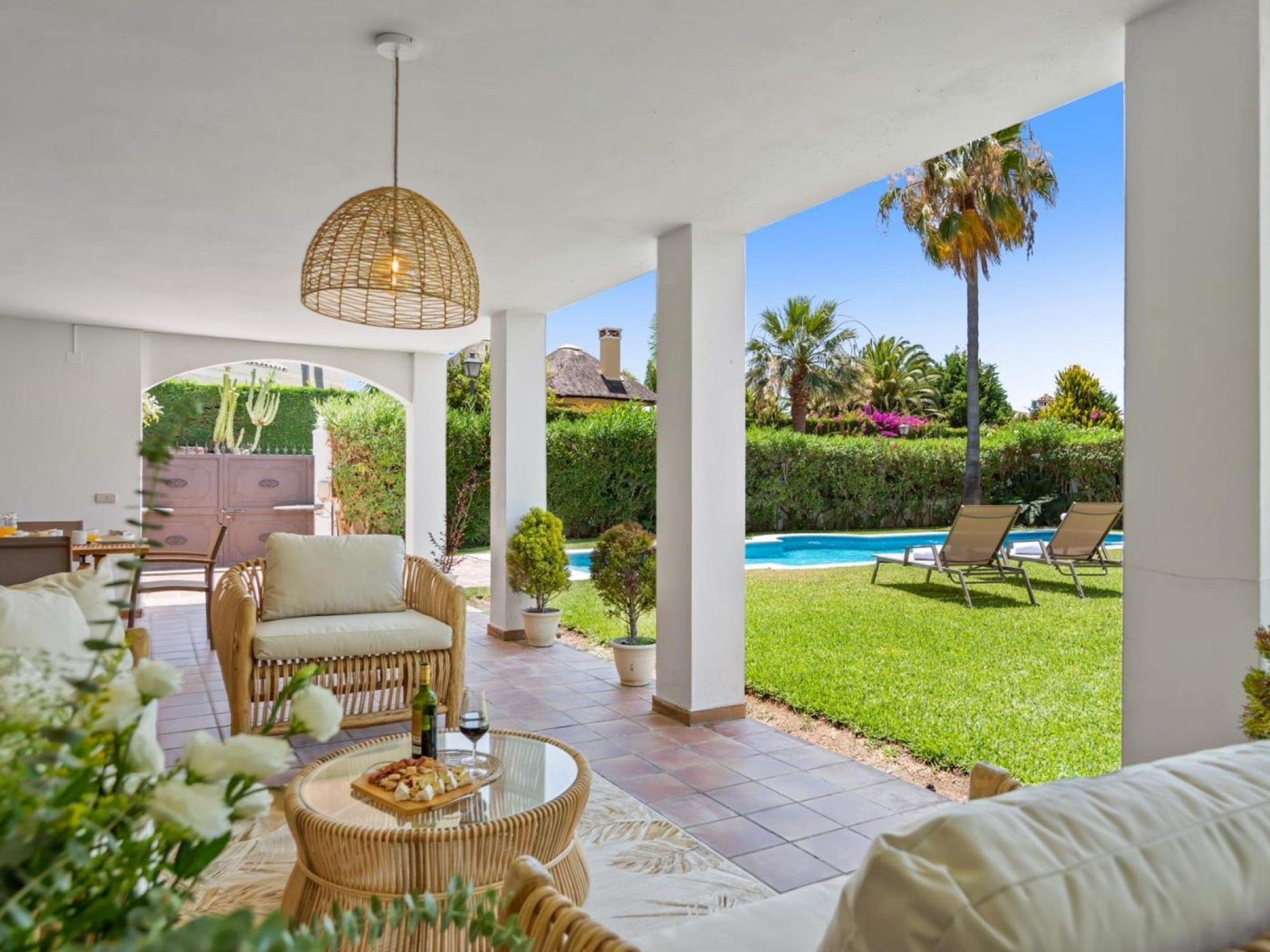 A covered outdoor seating area with wicker furniture, a glass table with food, and a view of a swimming pool and garden.