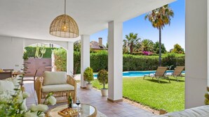 Ein überdachter Außenbereich mit Korbmöbeln, einem Glastisch mit Speisen und Blick auf Pool und Garten.