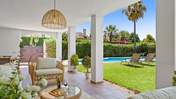 A covered outdoor seating area with wicker furniture, a glass table with food, and a view of a swimming pool and garden.